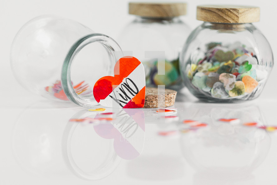 Jars of paper hearts on a white background with one lettered KIND in front.