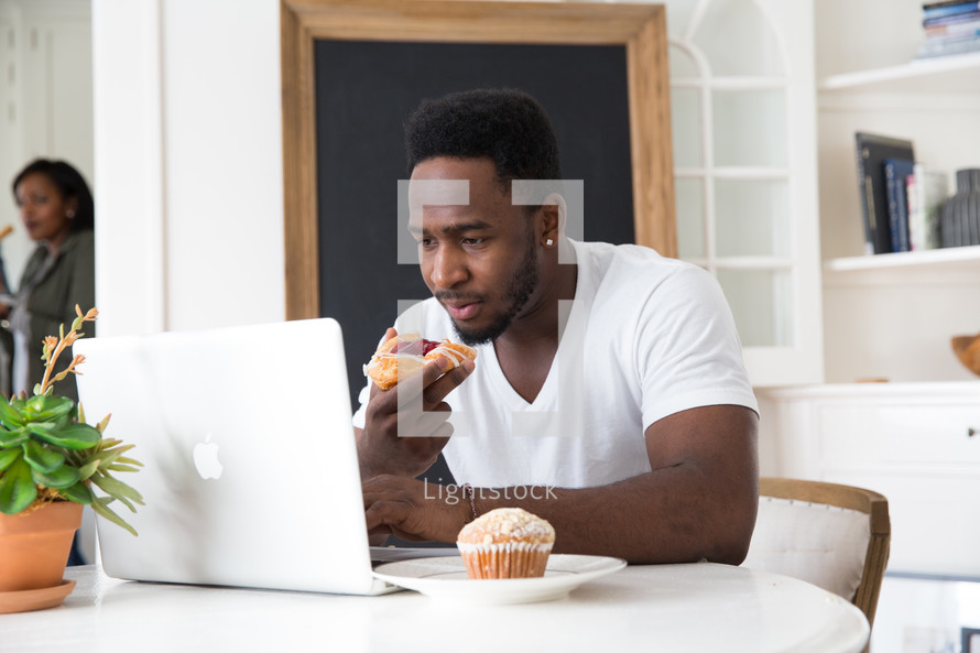 man eating a pastry while sitting in front of a laptop computer 