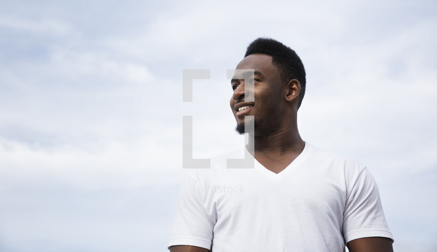 A smiling man in a white t-shirt against a cloudy white sky.