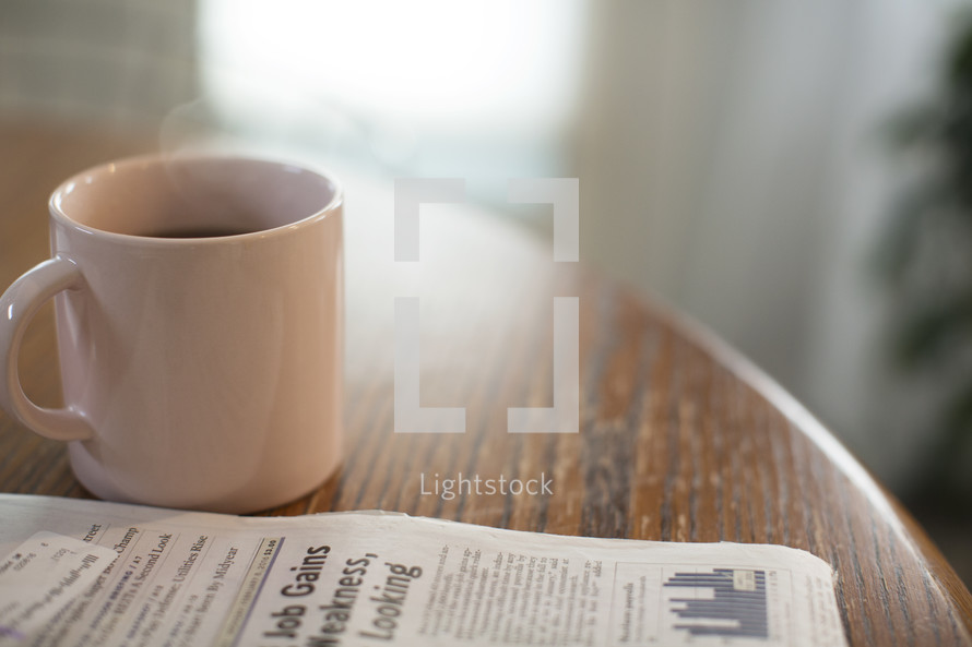 Morning light shining on a cup of coffee next to a newspaper on a table.