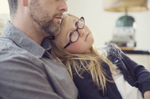 A little girl in glasses sits in her father's lap and looks at his face.