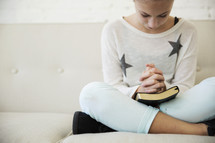a teen girl sitting on a couch praying over a Bible 