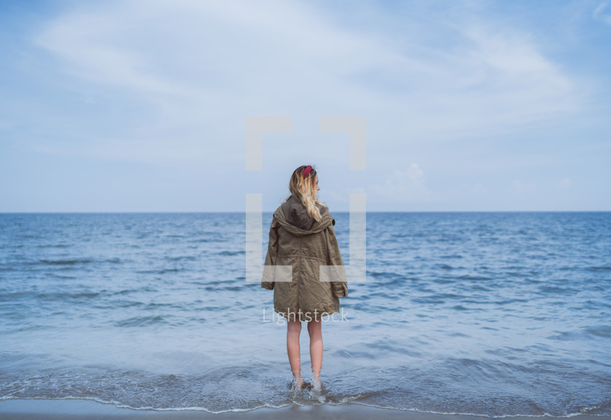 a girl in a coat standing in the ocean 