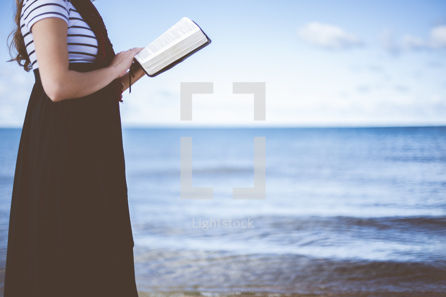 a woman standing on a beach reading a Bible 