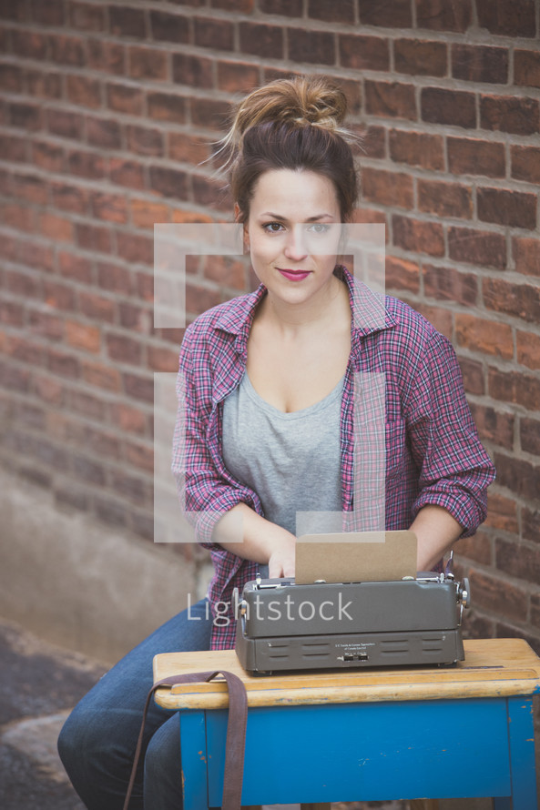 A young woman sitting at a school desk with a typewriter in front of a brick wall.