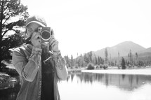 a woman taking a picture with a camera standing in front of a lake 
