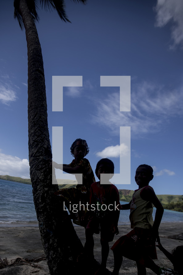 Silhouette of children leaning on a palm tree by the ocean.