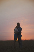 A man and woman hug in a field in front of a sunset.