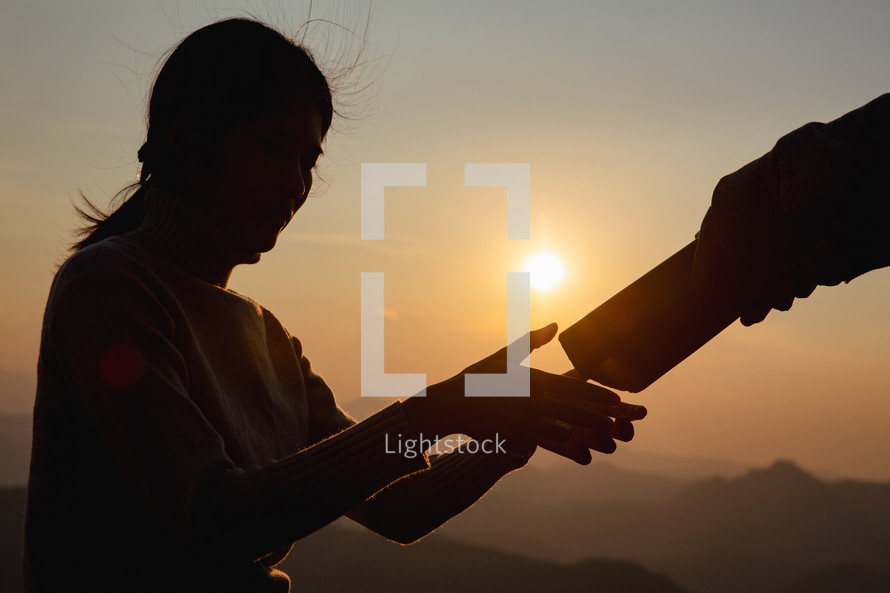 Silhouette of a man giving a Bible to a woman at sunrise.