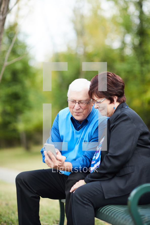 elderly couple sitting on a park bench looking at a cellphone 