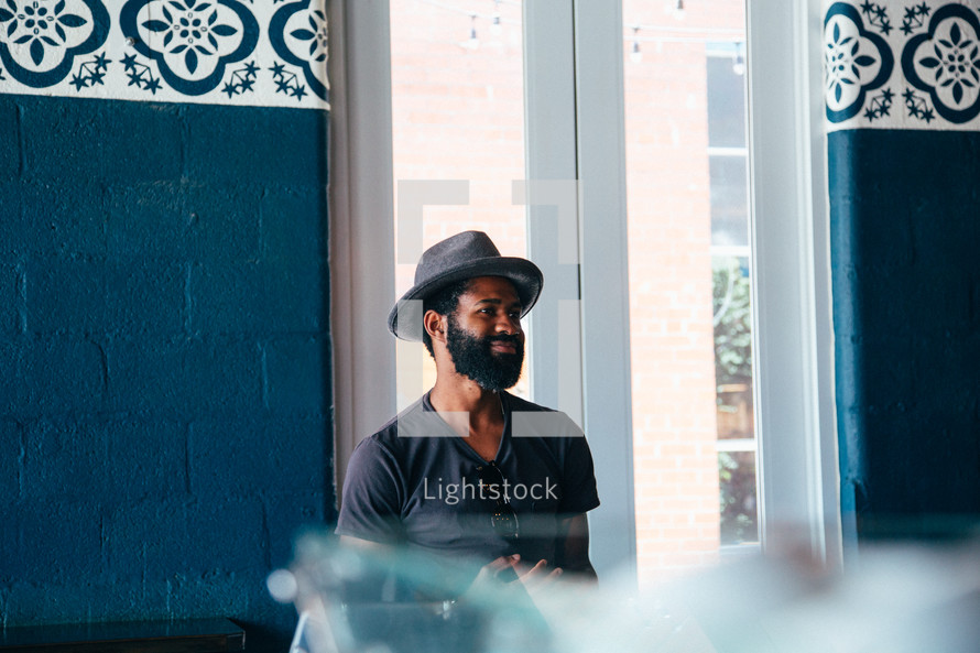 man in a hat and thick beard standing in front of a window 