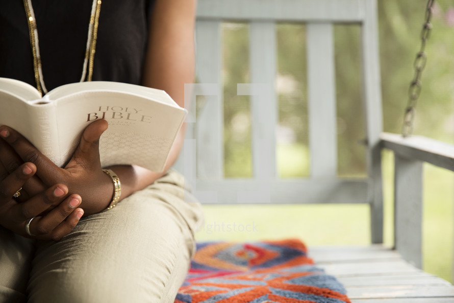 African-American woman sitting on a porch swing outside, reading a Bible.