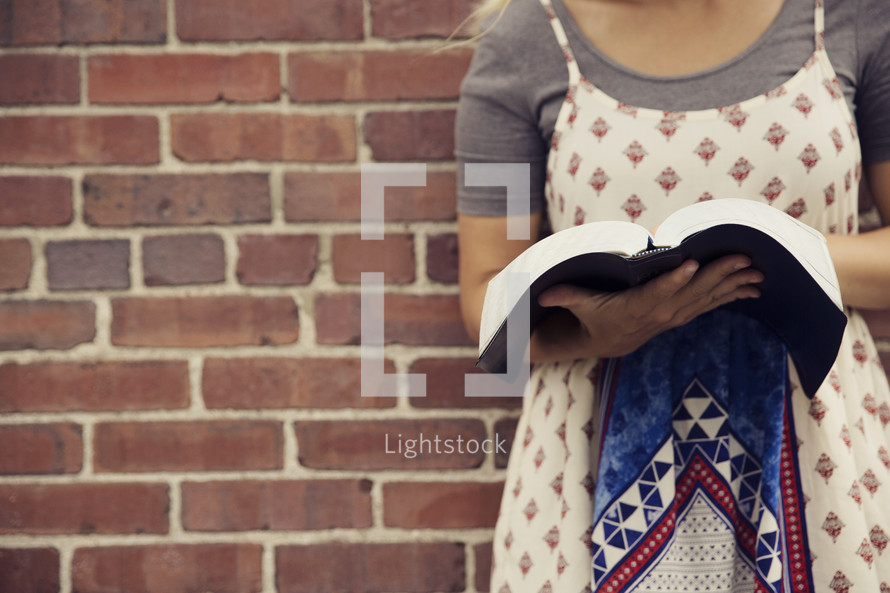 woman reading a Bible against a brick wall. 