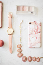 pink feminine watch, necklace, cellphone, makeup, and eyeshadow on a white countertop 