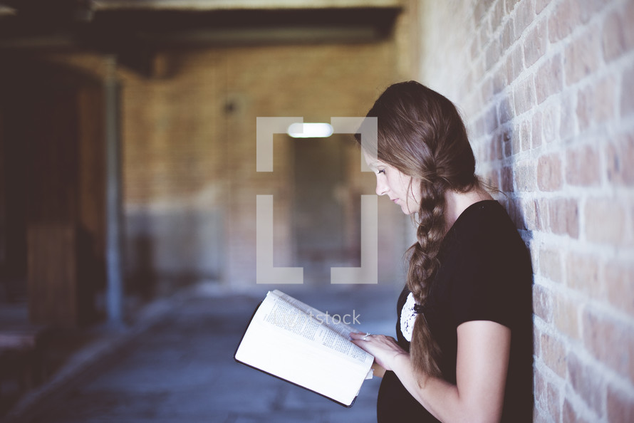 a woman leaning against a brick wall reading a Bible 