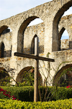 An old wooden cross stands in the stone courtyard and garden of a historic mission church, reminding visitors of the death and resurrection of Jesus Christ, along with the new life to be found in believing in Him.  