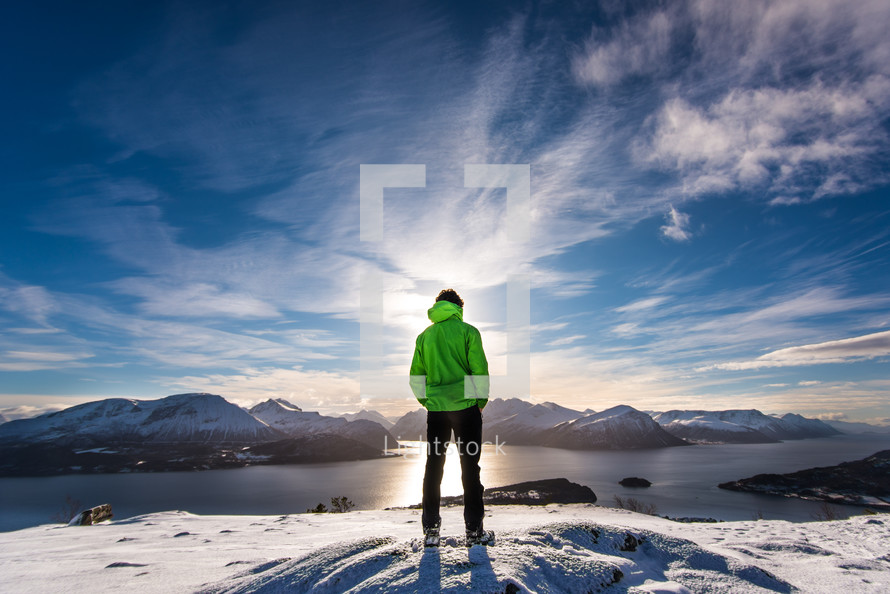 man standing on a snowy mountain looking out at a lake 