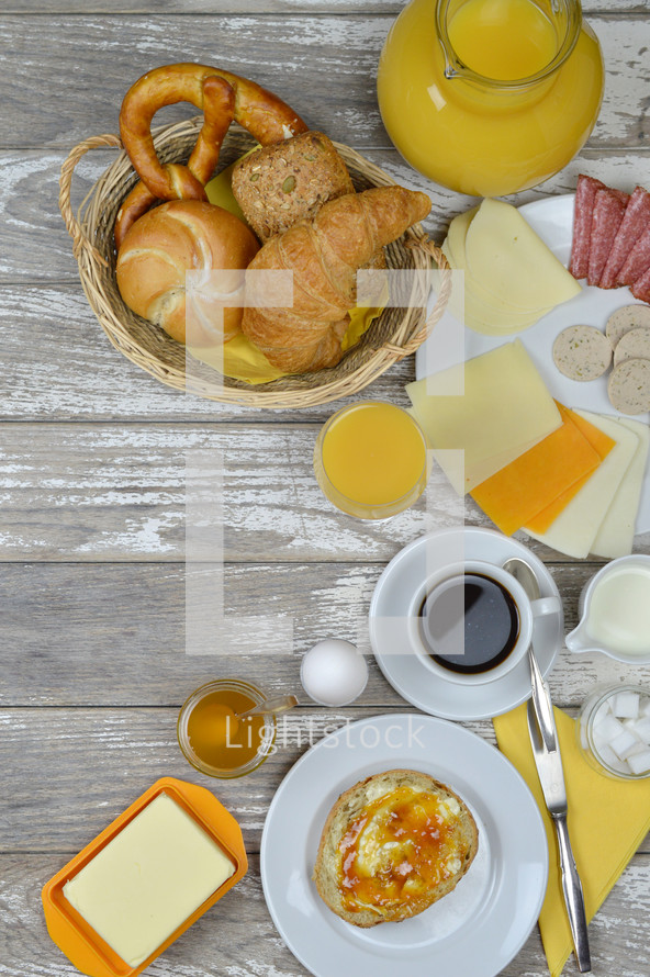 breakfast table with lots of fresh food like coffee, rolls, cheese, sausages, eggs, orange juice, jam, butter and a basket full of croissant, rolls and pretzel with copy space to the left