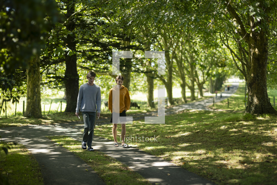 two young men walking and talking together on a driveway lined with trees in a beautiful setting