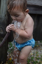 a little boy in a swim diaper drinking water from a hose 