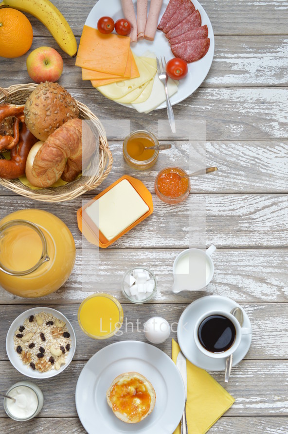 breakfast table with lots of fresh food like coffee, rolls, cheese, eggs, orange juice, tea, jam, butter, banana, apple, orange and a basket full of croissant, rolls and pretzel with copy space to the right