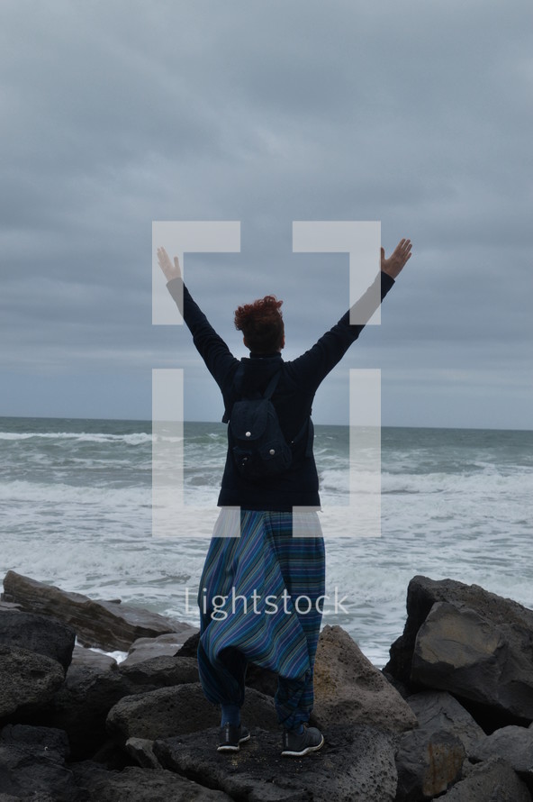 a woman with raised hands standing on a rocky shore 