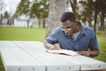 a man reading a Bible sitting at a picnic table in a park 