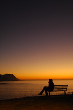 silhouette of a man sitting on a bench by a shore at sunset 