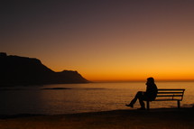 silhouette sitting on a bench by a shore at sunset 