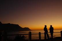 silhouettes of a people standing by railings along a shore at sunset 
