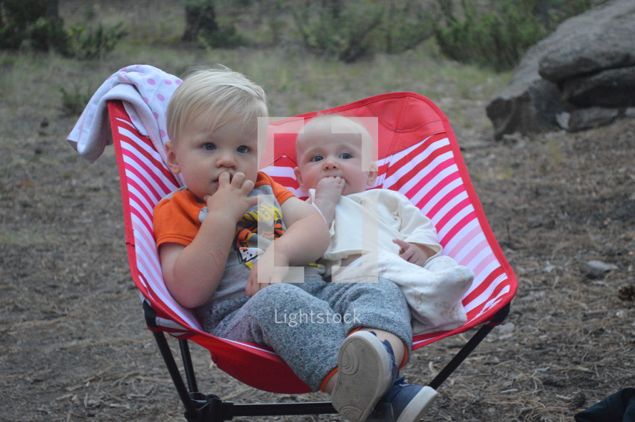 toddler brother and infant sister sitting together in a chair outdoors 