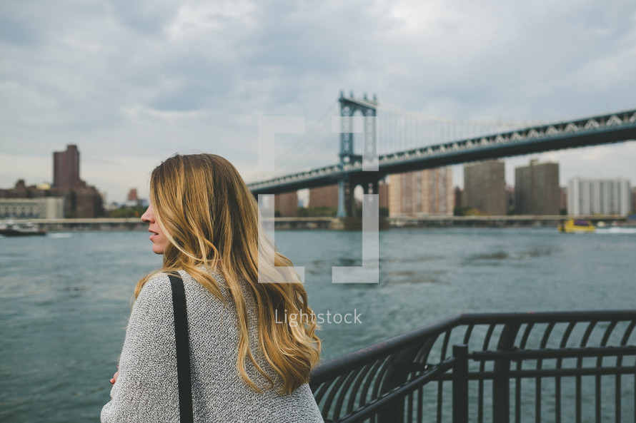 A woman stands at a railing looking out over a bay near a bridge.