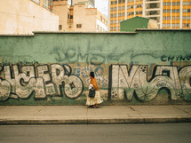 A Latin American woman walks along a graffiti covered wall.
