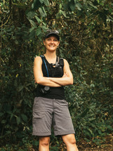 A smiling woman stands with her arms crossed against a background of trees and bushes.