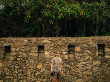 Man stands near an ancient stone wall among trees.