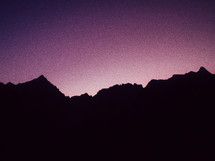 A purple sky illuminates a silhouette of mountains at dusk.
