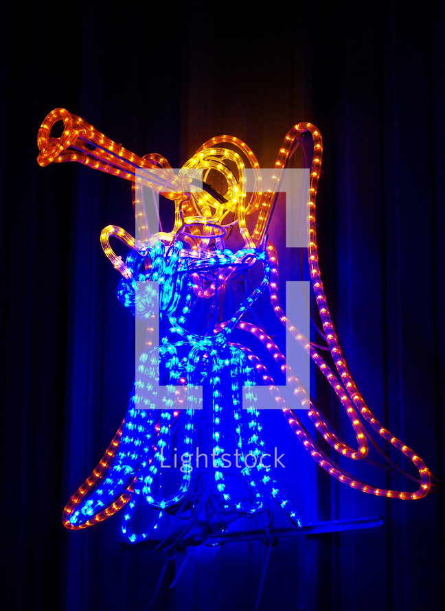 A Christmas Angel blows a trumpet announcing the birth of Jesus made out of neon blue and orange rope lights lighting up the night sky at Christmas with a blue and orange glow. 