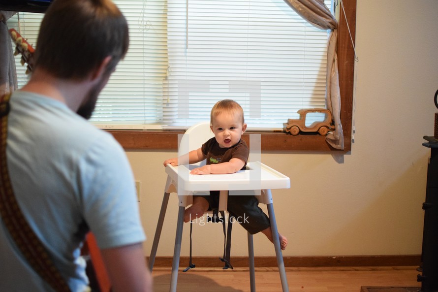 a father playing a guitar to a baby in a highchair 