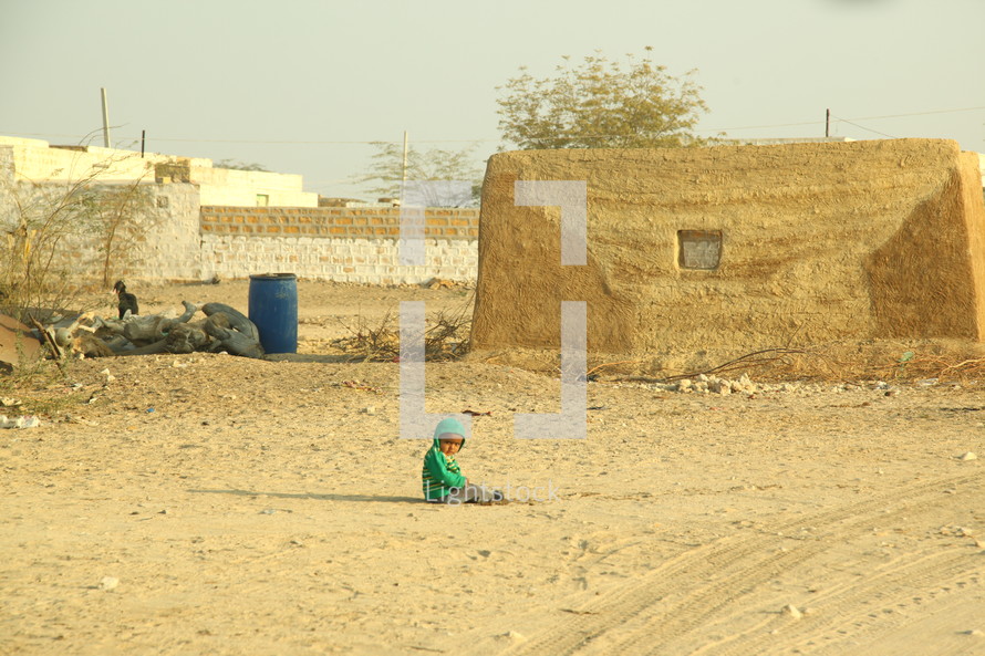 toddler sitting in sand in front of a stone hut 