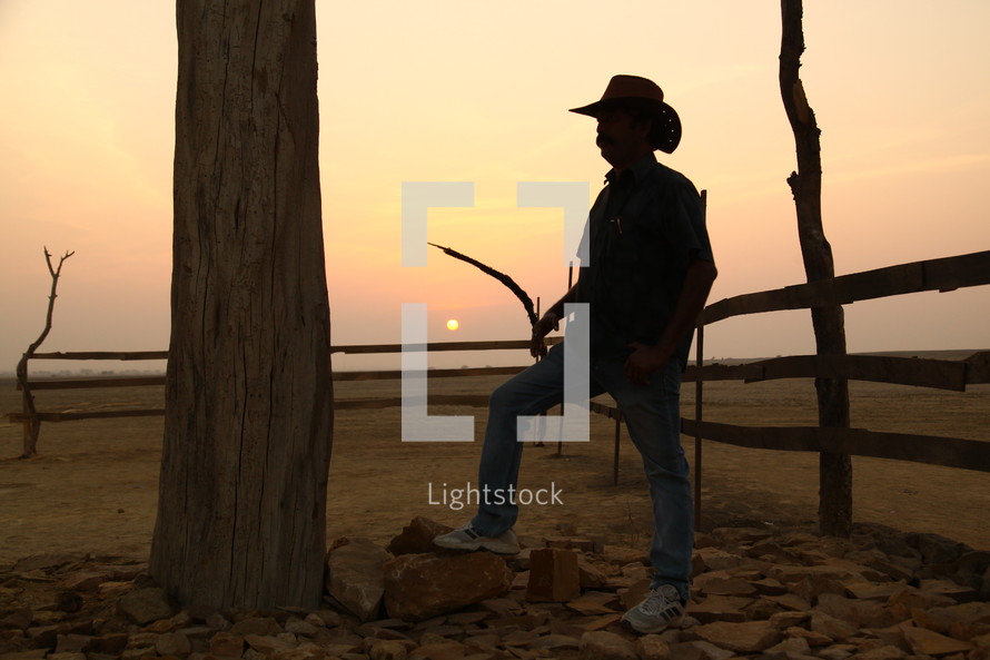 man in a cowboy hat standing by a fence in a desert 