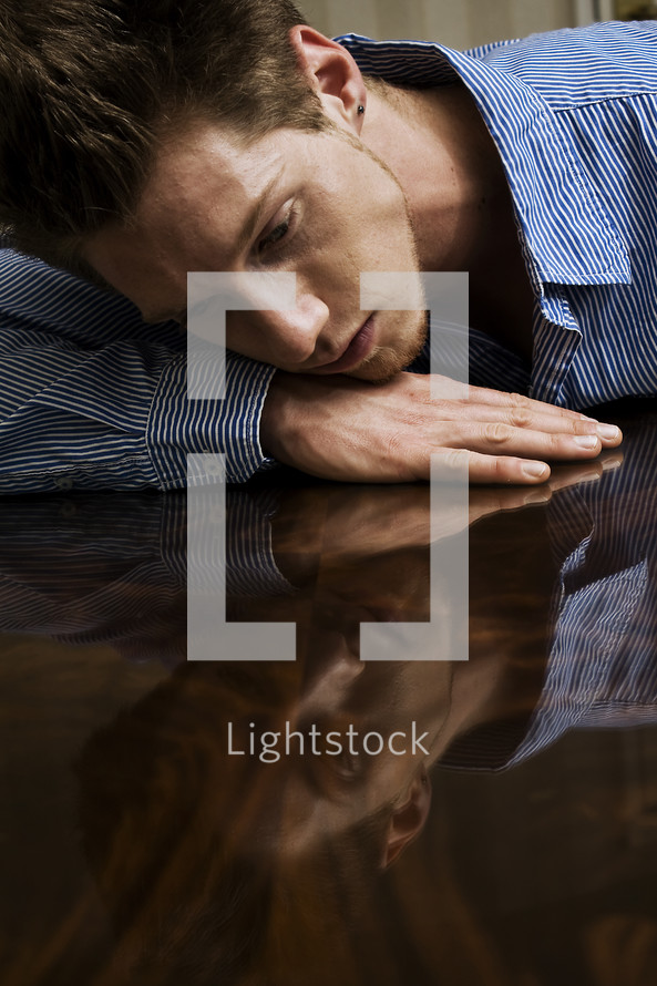 Man resting his head on hand lying on wood table top looking at reflection of his face.