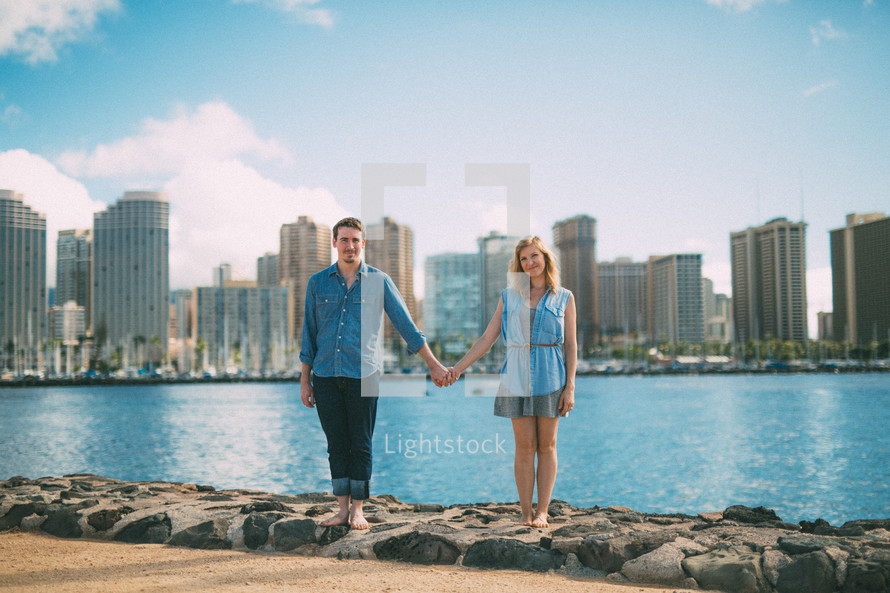 Couple holding hands on the river's edge with a city skyline on the background.