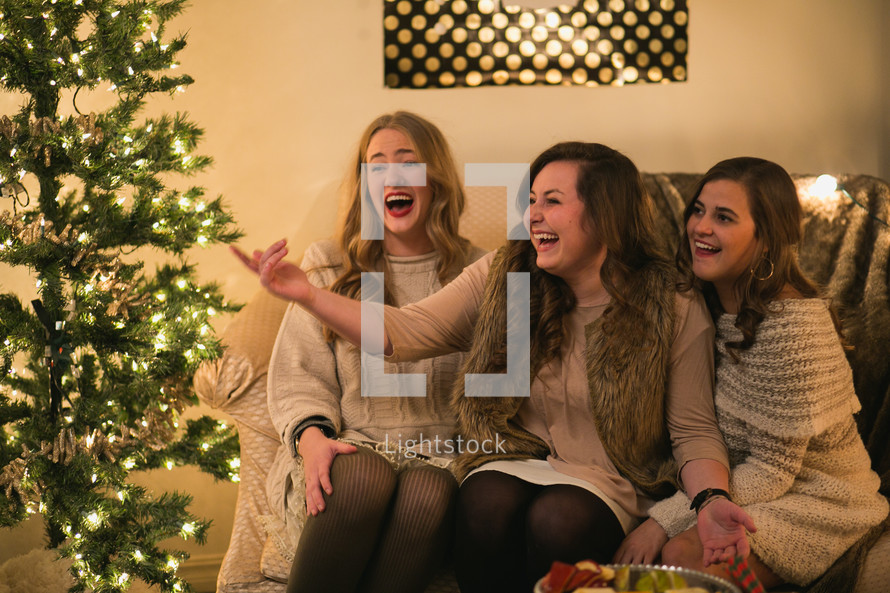Three laughing women sit together near a Christmas tree.