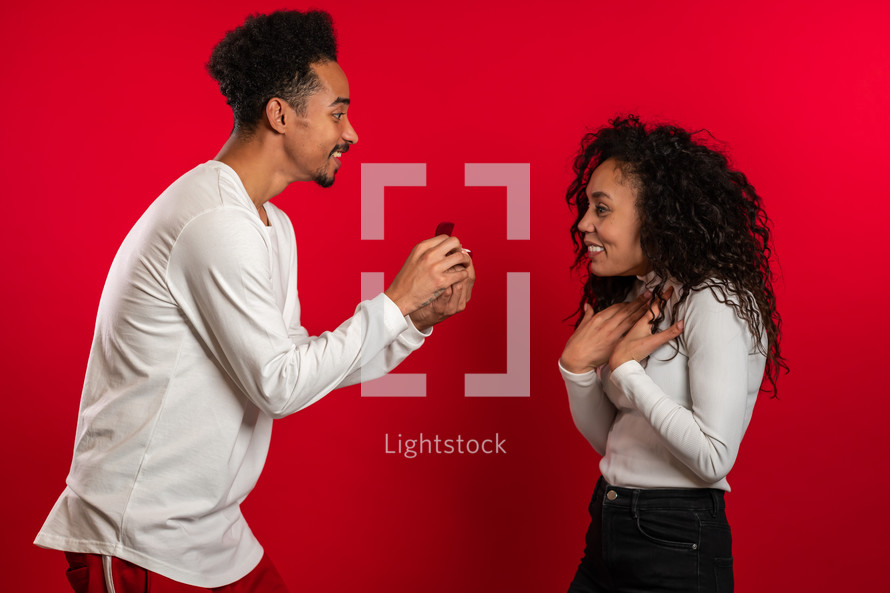 Young couple. African man makes marriage proposal to his lover woman with ring on red studio background. Love, holidays, happiness concept