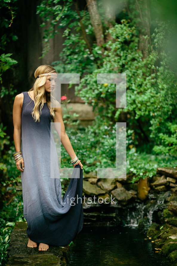a young blonde woman with a headband and long dress standing next to a koi pond 