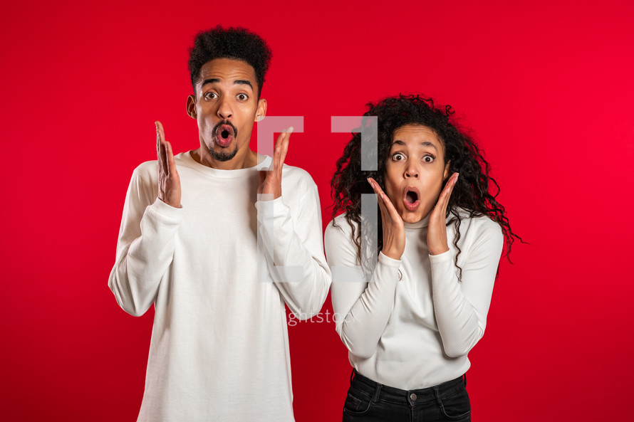 Amazed african american man and woman shocked, saying WOW. Young couple in white surprised to camera over red background.
