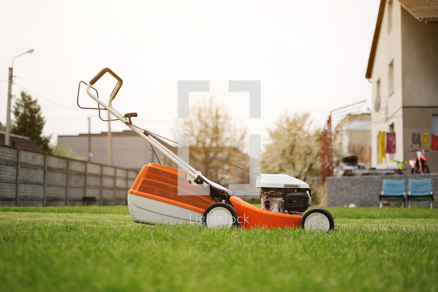Modern orange-grey electric lawn mower on bright lush green lawn at residential backyard of house. Gardening work tools. Rotary lawn mower machine cut grass. Professional lawn care service