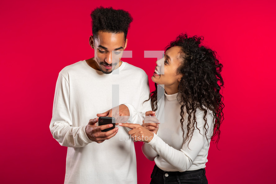 Young couple using mobile device. Man talking about apps and explains to girl how to use application. Red studio background.