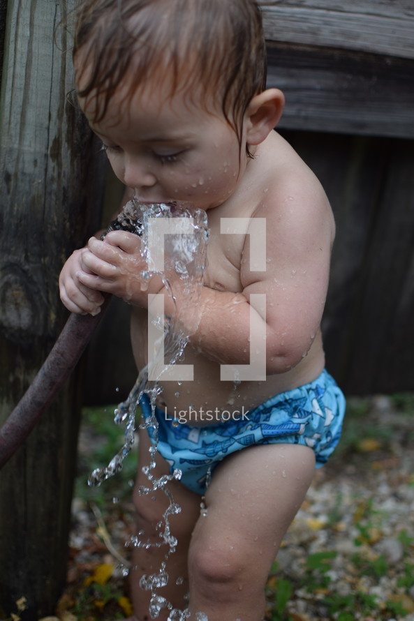 a little boy in a swim diaper drinking water from a hose 
