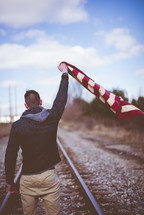 man standing on train tracks holding an American flag 